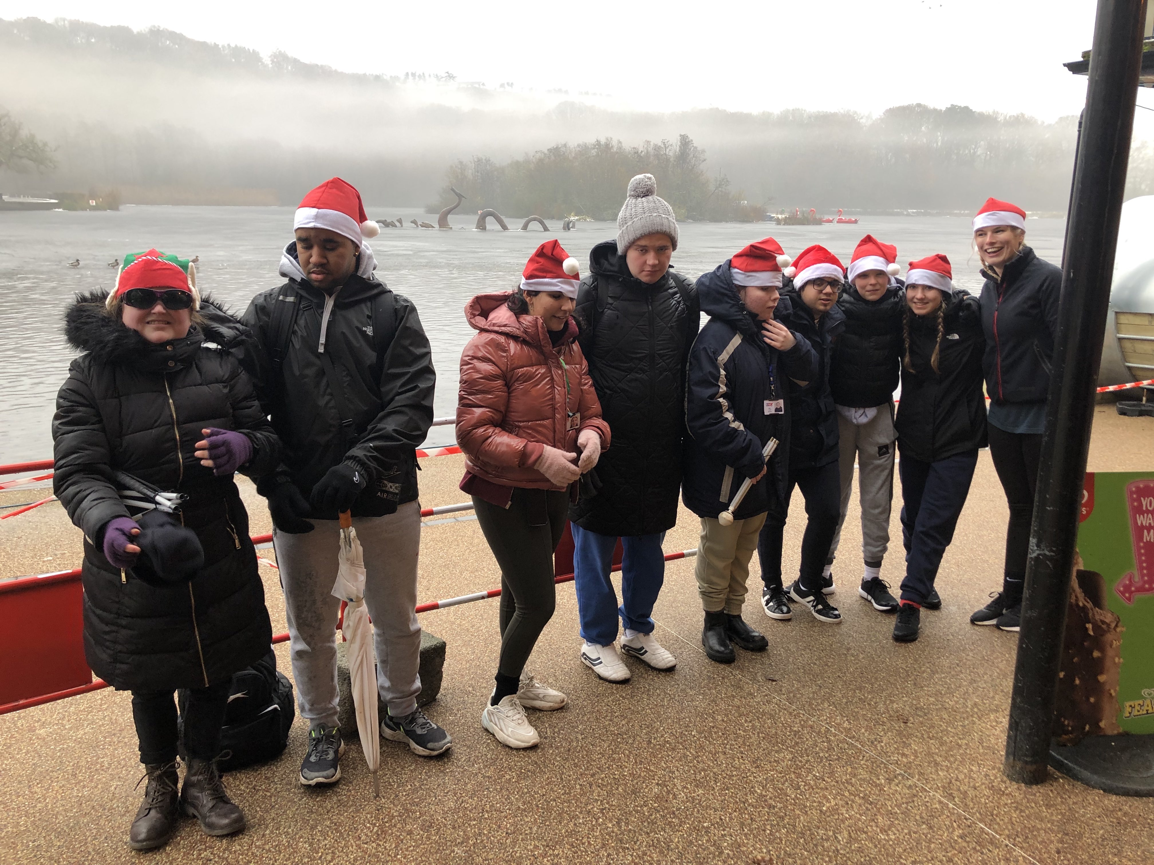 The students stand at the side of the lake wearing Santa hats and smiling