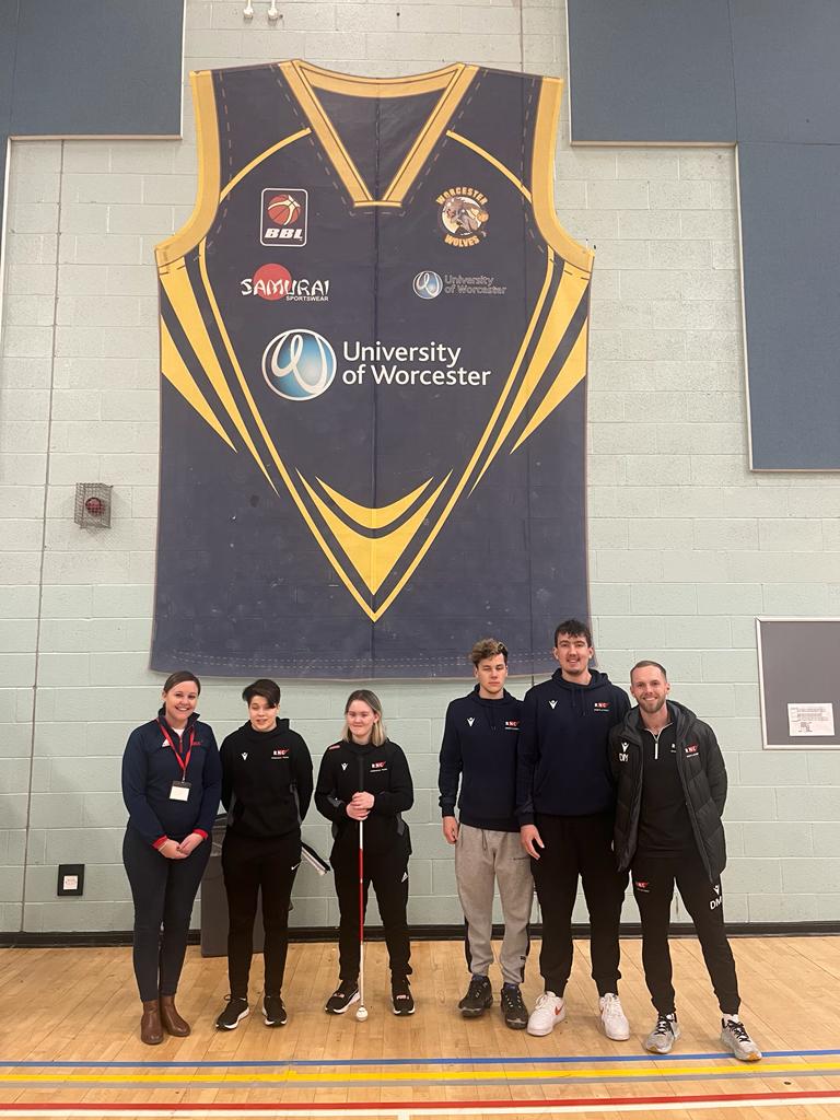 A group of five students with their tutor stand in front of a gigantic baseketball jersey on the wall in the University of Worcester sports hall