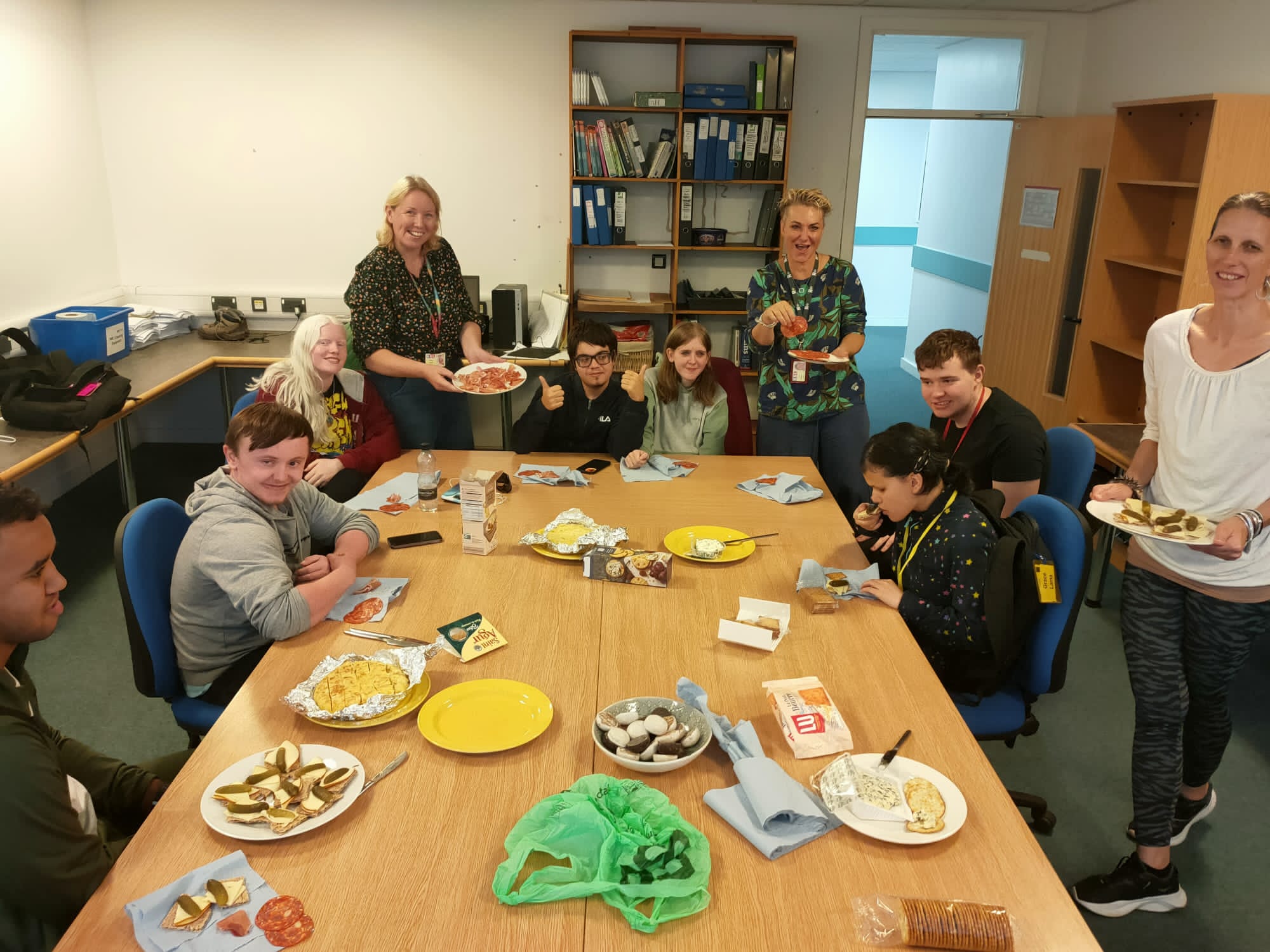 Students and staff sitting and standing around a table with lots of plates of European food to taste